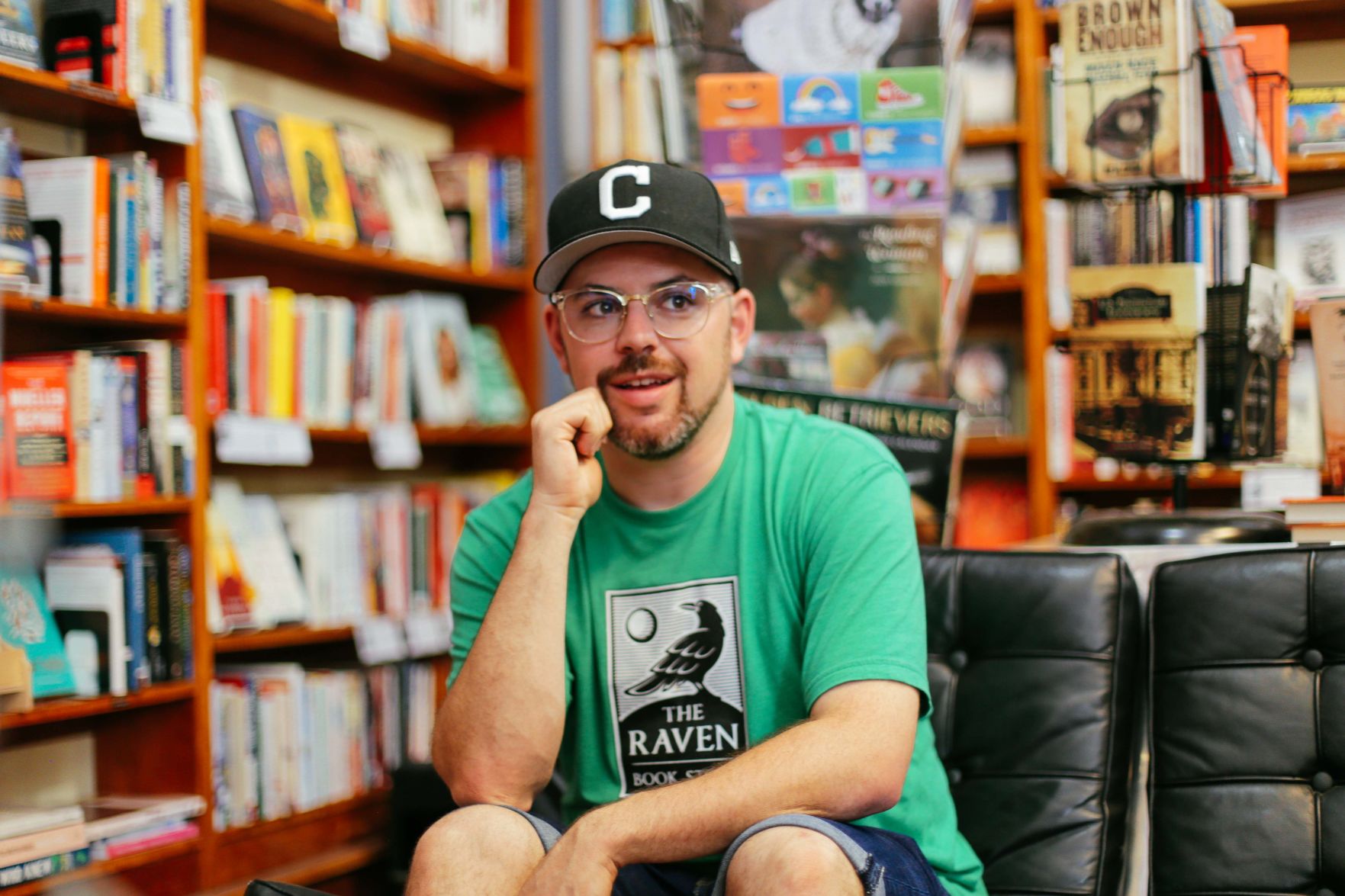 Danny Caine sits with his right fist holding his chin amid the bookshelves of The Raven Book Store